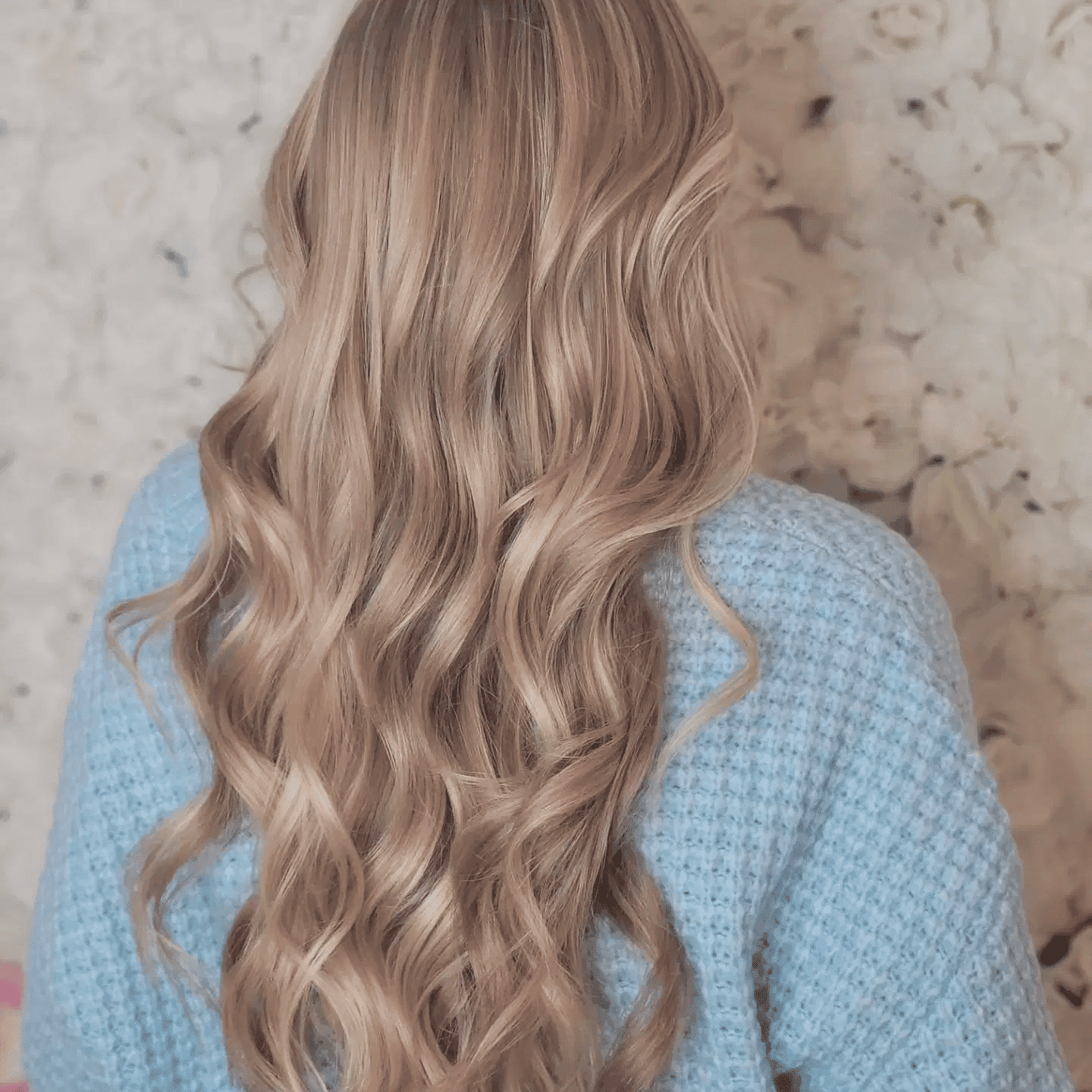 Woman with long, wavy blonde hair styled elegantly, wearing a blue knitted sweater, back view.
